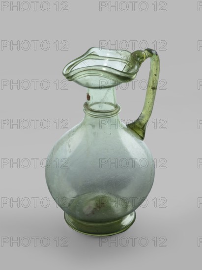 Oinochoe. Transparent, light green glass. Spherical belly on a foot with a clearance, created by pressing inward the central part of the bottom of the vessel; decoration on the cylindrical neck in the form of a single glass thread of the same color, wound horizontally. The vice is glued to the arms and extends in an arc to the edge of the trefoil rim; free-blown. 4th century, Cyprus; vessel oinochoe. Base diameter 4.8 cm; weight 0.062 kg., A translucent glass pitcher with a rounded body and a flared, undulating rim sits against a neutral background; the glass shows a pale green tint with areas of clearer and slightly darker green, a curved handle in a deeper green attached from the rim to the shoulder, a decorative ring around the neck, a short cylindrical neck leading to the wavy spout, and a circular foot at the base, with visible surface blemishes and irregularities in the glass.