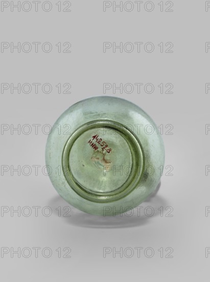 Oinochoe. Transparent, light green glass. Spherical belly on a foot with a clearance, created by pressing inward the central part of the bottom of the vessel; decoration on the cylindrical neck in the form of a single glass thread of the same color, wound horizontally. The handle is glued to the body and extends in an arc to the edge of the trefoil rim; free-blown. 4th century, Cyprus; vessel oinochoe. Base diameter 4.8 cm; weight 0.062 kg. Type: Isings form 124a., A round translucent glass object viewed from one end, showing a circular recessed base and a raised concentric ring, with a few irregularities and a small patch of residue on the inner recessed area; the glass is a pale green color with areas of lighter and darker green, and the background is a light gray.