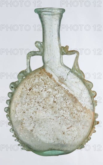 Ampoule, Egyptian workshop, 3rd century, Roman Empire 27 BC–476, vessel; height 12 cm, width 7.3 cm, A translucent glass vessel with a rounded, flattened body and a tall narrow neck, showing a pale green and clear glass color with areas of beige and brown surface encrustation; the body has a scalloped rim detail around its edges and two decorative looped handles near the neck that extend down onto the shoulder; the glass surface shows cloudy, pitted texture and a few visible hairline cracks.