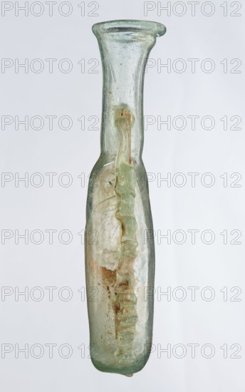 Ampoule, Egyptian workshop, 3rd century, Roman Empire 27 BC–476, vessel, height 12 cm, width 7.3 cm, A tall narrow glass vessel with a flared rim, shown upright against a light gray background, appears translucent pale green with areas of cloudy white and brown discoloration; a twisted ribbon of thicker green glass runs along the interior from near the neck down toward the base, creating a rippled pattern inside the body; the surface shows small bubbles and irregularities and a worn, slightly iridescent sheen, and the base is rounded with a small droplet-like protrusion.