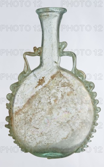 Ampoule, Egyptian workshop ; 3rd century, Roman Empire 27 BC–476; vessel; height 12 cm, width 7.3 cm, A translucent glass vessel with a rounded body and a narrow neck topped by a flared rim, showing a few irregular wavy handles attached near the neck and along the upper sides; the glass appears pale green with areas of cloudy white and iridescent sheens, and there are patches of brownish discoloration and surface weathering across the body; the edges of the body have a scalloped, ruffled profile and the surface shows small bubbles and streaks within the glass.