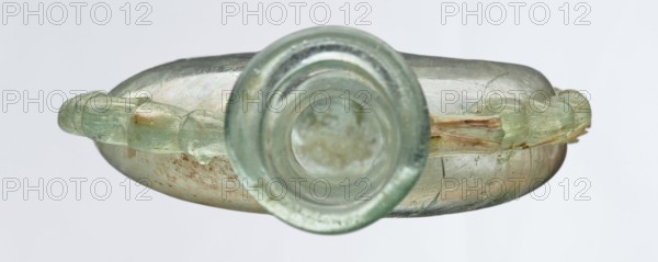 Ampoule, Egyptian workshop, 3rd century, Roman Empire 27 BC–476, vessel, height 12 cm, width 7.3 cm, A translucent glass vessel seen from above with a circular neck in the center and a rounded body extending outward, displaying a pale green and aquamarine hue with areas of iridescent sheen and cloudy discoloration; a decorative applied handle runs along each side, appearing as a group of linked rounded segments in a slightly greener shade with some amber-brown streaks and weathered surface texture; the body shows mottled patches of brownish residue and faint surface cracks, and the background is a neutral light gray.