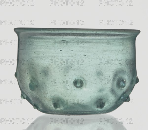 Bowl decorated with knobs; hemispherical shape with pinched projections;, 3rd–4th century AD. Isings form 96b2. Translucent green glass with visible streaks; free-blown. Outer rim diameter 9.30 cm; height 8.20 cm; outer bottom diameter 3.80 cm; weight 0.134 kg. Roman Empire 27 BC–476 origin., A translucent bowl-shaped vessel with a bluish-green color, shown against a pale gray background, with a rounded rim and a slightly flared lip; the glass surface has a few raised bumps and rounded protrusions around the lower half, small air bubble inclusions, and subtle surface striations and imperfections; the base reflects faintly on the surface below, and overall tones include teal, aqua, pale green, gray, and hints of darker bluish-green.