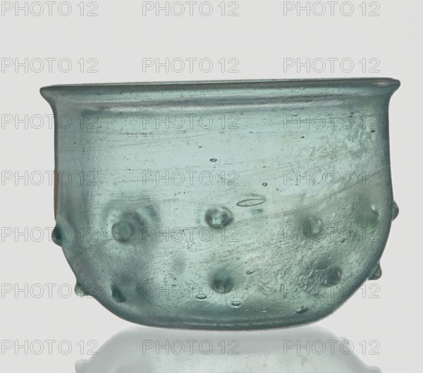 Bowl decorated with knobs; hemispherical shape with pinched projections; maker, 3rd–4th century AD. Type: Isings form 96b2. Translucent green glass with visible streaks in the glass mass; free-blown. Outer diameter of rim: 9.30 cm; height: 8.20 cm; outer diameter of bottom: 3.80 cm; weight: 0.134 kg. Origin: Roman Empire 27 BC–476., A translucent glass vessel with a rounded shape and a flared rim, shown against a pale background; the glass is a bluish-green color with variations in tone and a few cloudy areas, and contains several small raised bubbles and irregularities on its surface; the base of the vessel is slightly flattened and reflects faintly on the surface beneath, and the overall surface shows fine scratches and texture.