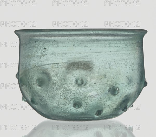 Bowl decorated with knobs. Hemispherical shape with pinched projections;, 3rd–4th century AD. Isings form 96b2. Translucent green glass with visible streaks; free-blown. Rim diameter 9.30 cm; height 8.20 cm; bottom diameter 3.80 cm; weight 0.134 kg. Roman Empire 27 BC–476., A translucent greenish-blue glass bowl with a rounded body and a slightly flared rim, showing a textured surface with small raised bumps and irregularities; the glass displays variations in color from pale aqua to deeper teal, with cloudy patches and streaks, and a glossy finish that reflects light against a pale neutral background.