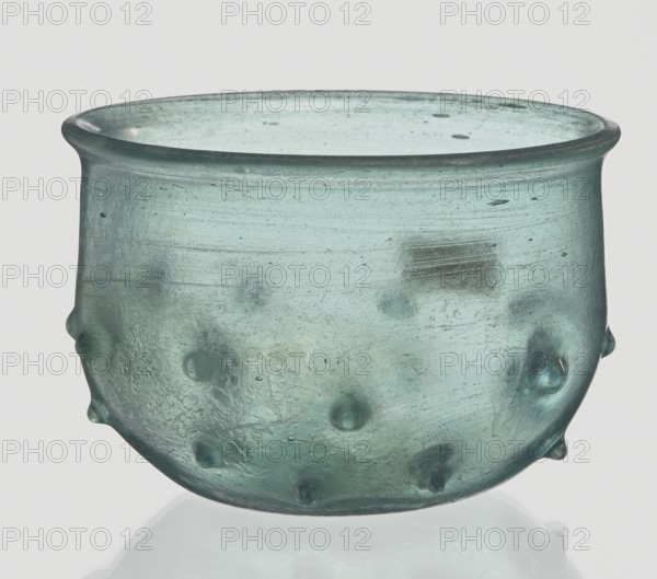 Bowl decorated with knobs; hemispherical shape with pinched projections;, 3rd–4th century AD; Isings form 96b2; translucent green glass with visible streaks; free-blown; outer rim diameter 9.30 cm; height 8.20 cm; outer bottom diameter 3.80 cm; Roman Empire 27 BC–476; vessel; weight 0.134 kg., A translucent bowl-shaped glass vessel with a rounded body and a slightly flared rim, shown against a pale background; the glass is a pale greenish-blue with areas of subtle variation in tone, cloudiness, and small dark specks, and the surface displays circular impressions and several rounded protrusions on the exterior; the rim is thicker and darker along its edge, and the interior shows faint horizontal striations and scattered small bubbles, while the base area curves inward and reflects the pale background.