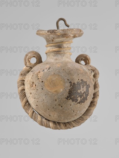 Ampoule, Egyptian workshop, 4th century BC, vessel, height 5.2 cm, width 4.2 cm, A rounded ceramic vessel with a narrow neck and flared rim, showing a textured surface with pitting and areas of darker wear, is suspended against a plain light gray background; the vessel has a pair of looped handles near the neck and a twisted rope-like adornment encircling its lower half, and a metal hook is inserted at the rim. The visible colors include beige, tan, light brown, darker brown patches, and gray.