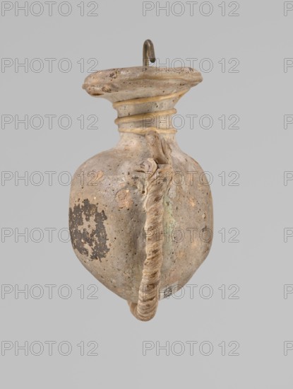Ampoule, Egyptian workshop, 4th century BC, vessel, height 5.2 cm, width 4.2 cm, An aged, bulbous ceramic vessel with a rounded body and a flared neck, shown against a plain light gray background; the vessel surface is mottled with shades of beige, tan, brown, and hints of pale green, and shows areas of darker brown patina and spots of weathering; a thick twisted handle runs from near the neck down the side to the base, the handle appearing textured and beige with darker accents; the rim is irregular and slightly worn, with concentric ridges at the neck that are highlighted in a pale tan; a small metal hook protrudes from the top of the image, showing a dark brown, slightly corroded surface.
