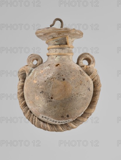 Ampoule, Egyptian workshop, 4th century BC, vessel, height 5.2 cm, width 4.2 cm, A rounded ceramic vessel displayed against a plain gray background, showing a bulbous body with a narrow neck and a flared rim; the surface of the vessel is mottled with beige, tan, light brown, and small darker brown speckles, with areas of lighter cream and pale gray; two looped handles rise from the shoulder, each with a coiled rope-like attachment that curves down and around the base, the rope-like element appearing in similar beige and tan tones with shadowed ridges; the rim has a hooked metal loop inserted, which is dark brown with a slightly rusty patina; the vessel surface shows small pitted holes and textured wear in various spots, and a slightly darker circular area is visible on the front of the body.