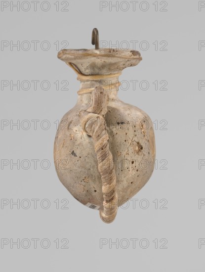 Ampoule, Egyptian workshop,  workshop, 4th century BC, vessel, height 5.2 cm, width 4.2 cm, An aged ceramic vessel with a rounded body and a narrow neck topped by a flared rim, shown against a plain light gray background; the surface of the vessel is mottled with beige, tan, light brown, and gray tones with darker brown speckles and small weathered patches; a twisted rope-like handle runs from the neck to the lower body, matching the vessel's beige and tan coloration with visible texture; the rim has a slightly darker beige band and a small metal hook projecting upward from its center; the vessel shows signs of wear, small pits, and irregularities in color and surface texture.