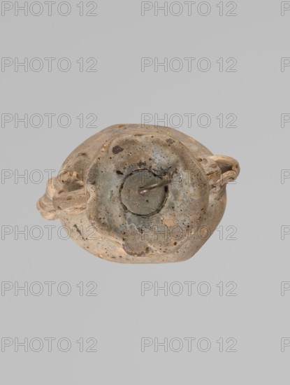 Ampoule, Egyptian workshop, 4th century BC, vessel; height 5.2 cm, width 4.2 cm, A worn, round object with a rough surface in shades of beige, tan, gray, and brown sits against a flat gray background; the object has an irregular rim and a central circular depression with a darker gray-green area and a small protruding piece, and a pair of sculpted handles or looped extensions on opposite sides showing the same beige and tan tones with darker spots and speckles.