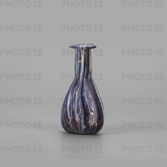 Unguentarium, unknown eastern workshop, translucent glass, mosaic: blue-violet-white. Pear-shaped body, cylindrical neck, rim tilted outwards and curved inwards, flattened; sagging/free blowing. End of 1st century BC–first half of 1st century AD. Unguentarium vessel, rim diameter 2.3 cm, base diameter 2.05 cm, weight 0.030 kg. Type: Isings form 6/28., A single small glass vessel with a narrow neck and flared lip sits on a flat surface against a plain background; the vessel has a bulbous base tapering into a slender neck and displays a glossy finish with swirling vertical streaks in shades of dark blue, deep purple, pale pink, and touches of gold, along with a few lighter oval spots and subtle surface imperfections.