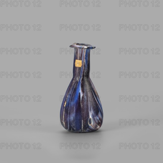 Unguentarium, unknown eastern workshop, translucent glass mosaic blue-violet-white. Pear-shaped body, cylindrical neck, rim tilted outwards and curved inwards, flattened; sagging/free blowing. End of 1st century BC–first half of 1st century. Vessel unguentarium. Rim diameter 2.3 cm; base diameter 2.05 cm; weight 0.030 kg. Type: Isings form 6/28., A single small vase with a narrow flared rim and a bulbous body sits against a plain background, showing a glossy surface with swirling bands of deep blue, lighter blue, white, and hints of purple, along with a small area of yellowed paper stuck near the neck; the vase has vertical ribbing, a slightly irregular rim, and a reflective sheen with some worn spots and small imperfections.