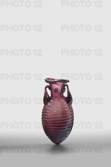 Amphora, unknown eastern workshop; De Tommaso form 20. Translucent manganese glass with an elongated ovoid body covered with horizontal ribs and an outwardly flaring rim; body blown in a two-part form, neck and rim freely blown. Outer rim diameter 3 cm; 2nd half of the 1st century. Vessel amphora, weight 0.024 kg., A small ribbed vase with a rounded, elongated body and a flared, irregular rim, shown against a plain light gray background; the vase is primarily purple with gradients of darker purple and hints of reddish tones, and it has a pair of curved handles on either side that appear darker in color, almost brownish-purple, with subtle glossy reflections and some areas of lighter wear.