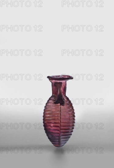 Amphora, unknown eastern workshop; glass vessels, Type: De Tommaso form 20. Translucent manganese glass; elongated ovoid body with horizontal ribs and an outwardly flaring rim; body blown in a two-part form, neck and rim freely blown. Outer diameter of rim: 3 cm; 2nd half of the 1st century; place of origin, vessel amphora; weight 0.024 kg., A small translucent vessel with a ribbed, elongated body and a narrow neck topped by a flared rim, shown against a plain light gray background. The object is primarily a deep purple to reddish-purple color with variations in tone that become slightly darker toward the bottom and around the rim. The surface shows horizontal ridges encircling the body and a vertical seam or central line running along its length. A short, irregular drip or applied piece of glass projects from the neck area, darker in color than the surrounding glass. The lighting creates subtle highlights and soft shadows beneath the vessel, and the overall scene includes shades of gr...