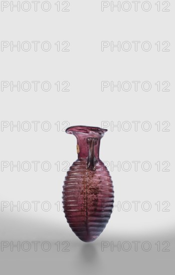 Amphora, unknown eastern workshop; Translucent, manganese glass; elongated ovoid body with horizontal ribs and an outwardly flaring rim; body blown in a two-part form, neck and rim freely blown; outer diameter of the rim 3 cm; 2nd half of the 1st century; vessel amphora; weight 0.024 kg., A ribbed, elongated glass vessel with an ovate body and a flared, irregular rim sits against a pale background, showing a translucent purple hue with variations from deep plum to lighter pinkish tones and areas of brownish clouding; the vessel has a narrow neck with a small attached handle-like feature in a darker purple and some surface weathering and texture, casting a soft shadow beneath it.