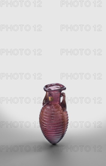 Amphora, unknown eastern workshop; translucent manganese glass; elongated ovoid body covered with horizontal ribs and an outwardly flaring rim; body blown in a two-part form, neck and rim freely blown. Outer diameter of the rim: 3 cm; 2nd half of the 1st century; vessel, amphora; weight 0.024 kg., A small translucent vessel with a ribbed, rounded body and a narrow neck topped by a flared lip, shown against a pale gray background; the vessel has a single curved handle attached near the neck, and its surface shows areas of rough texture and wear; the visible colors are shades of purple and pink with hints of brown and darker areas.