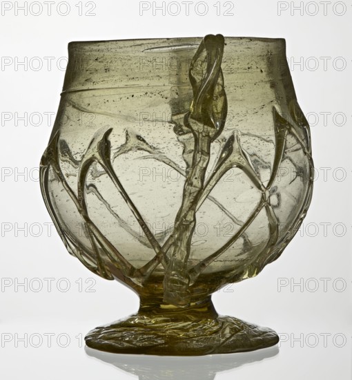 Skyphos, deep bowl with wide foot and two small vertical handles. Surface decorated with a zigzag ornament applied with irregular threads. 3rd–4th century AD, Roman Empire 27 BC–476. Vessel, height 11 cm., A translucent glass vessel with a rounded bowl and a short stem on a flattened foot, primarily pale olive-green in color with darker green accents; the surface shows irregular bubbles and streaks, and several raised twisted glass ribbons and arc-shaped applied elements wrap around the bowl creating a decorative network; the rim is slightly uneven and the base appears textured with rippled glass, all shown against a light neutral background.