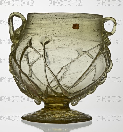 Skyphos, deep bowl with wide foot and two small vertical handles. Surface decorated with a zigzag ornament applied with irregular threads. 3rd–4th century AD, Roman Empire 27 BC–476. Vessel, height 11 cm, A translucent glass vessel with a rounded body on a flattened foot, featuring two looped handles on opposite sides and raised applied trails forming looping and branching decorations across the surface; the glass appears pale greenish-yellow with darker olive-green areas at the foot and along some applied trails, and there are a few small bubbles and inclusions visible within the glass.