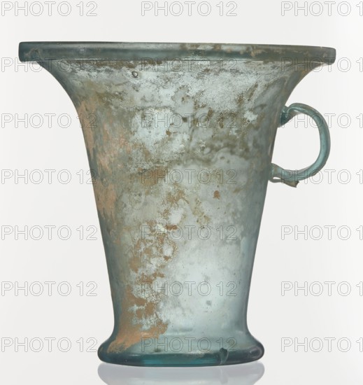 Modiolus – drinking vessel, Goluchów Greater Poland Voivodeship, 1st century, Roman Empire 27 BC–476, vessel, height 14.4 cm, width 15.4 cm, A translucent glass vessel shaped like a flared cup with a rounded base and a single loop handle on one side, showing mottled surface areas of greenish-blue and pale aqua with patches of brownish-gold weathering, a smooth rim and a slightly glossy finish, photographed against a pale neutral background.