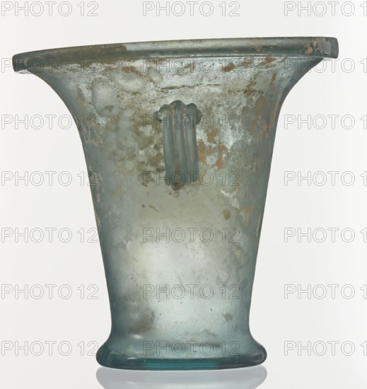 Modiolus – drinking vessel, Goluchów Greater Poland Voivodeship, 1st century, Roman Empire 27 BC–476, vessel, height 14.4 cm, width 15.4 cm, A single glass vessel shown upright with a flared rim and a rounded foot, rendered in translucent bluish-green glass with areas of cloudiness and irregular surface texture; patches of brownish discoloration and pale whitish film appear across the body, and a small applied loop handle of the same bluish-green glass is attached near the upper middle; lighting creates subtle highlights and reflections on the glass surface and a faint reflection of the base on the white background.