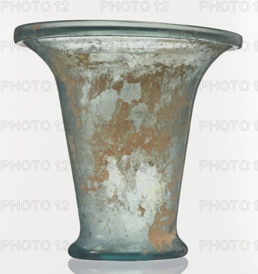 Modiolus – drinking vessel, Goluchów Greater Poland Voivodeship, 1st century, Roman Empire 27 BC–476, vessel, height 14.4 cm, width 15.4 cm, A single large flared vessel with a broad rim and a tapered base sits against a plain light background, showing a weathered surface with patches of bluish-green glass intermingled with areas of brownish corrosion and paler gray-green encrustation; the rim and base edges appear slightly darker teal, and the vessel displays irregular mottled textures and worn spots across its body.