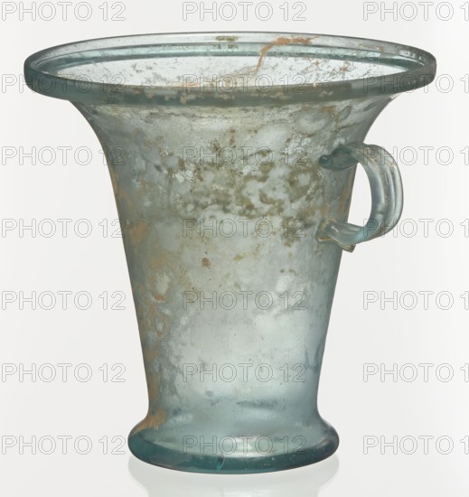 Modiolus – drinking vessel, Goluchów Greater Poland Voivodeship, 1st century, Roman Empire 27 BC–476, vessel, height 14.4 cm, width 15.4 cm, A translucent glass vessel with a flared rim and a rounded base, showing a weathered, mottled surface with a mix of pale blue, aqua, green, gray, and areas of brownish discoloration; it has a single looped handle attached near the upper side, the handle sharing the pale blue and aqua tones, and the glass shows bubbles, cloudiness, and surface irregularities across its body.