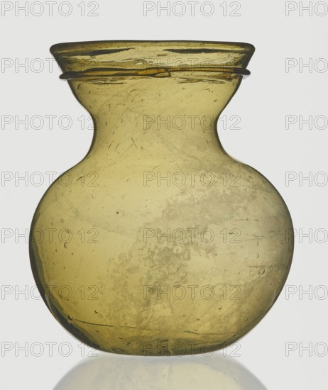 A viceless vessel, Goluchów Greater Poland Voivodeship, below the edge a glass thread is applied horizontally. Unspecified place of origin, vessel, height 12.4 cm, width 10.7 cm, A translucent rounded glass vessel with a narrow neck and flared rim, shown against a light background; the glass is an olive-greenish yellow with variations in tone, areas of cloudiness and internal bubbles, and a few darker green streaks and speckles visible throughout the surface, with a subtle reflection beneath it.