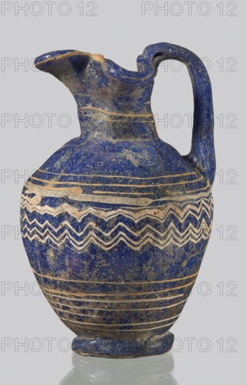 Oinochoe with a vertical arch-shaped vice; eastern workshop;, 3rd century BC. Type: Grose group II:A, form II:3. Dark blue glass; on the belly threaded decoration in the form of horizontal and zigzag lines at the widest point of white and yellow glass. Trefoil rim; molding on the core. Height: 8.90 cm. Body diameter: 5.60 cm. Outer diameter of the rim: ? cm. Outer diameter of the bottom: ? cm. Vessel oinochoe. Weight: 0.076 kg., A blue ceramic jug with a rounded body, narrow neck, curved spout with a chipped rim, and a single arched handle; the surface shows a deep cobalt blue with lighter blue speckling and areas of worn beige where the glaze has flaked, and a decorative band of zigzag and wavy patterns in beige encircling the midsection, along with several horizontal beige bands near the shoulder and base.