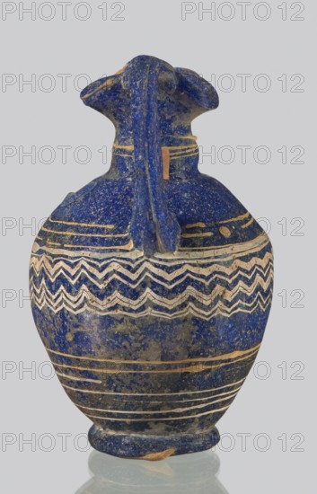 Oinochoe with a vertical arch-shaped vice; eastern workshop;, 3rd century BC. Type: Grose group II:A, form II:3. Dark blue glass with threaded decoration on the belly in horizontal and zigzag lines at the widest point of white and yellow glass. Trefoil rim; molding on the core. Height: 8.90 cm. Body diameter: 5.60 cm. Outer diameter of the rim: ? cm. Outer diameter of the bottom: ? cm. Vessel oinochoe. Weight: 0.076 kg., A small rounded vessel made of a speckled deep blue material with a glossy finish, featuring a narrow neck and a short flared rim with two rounded projections on opposite sides; the body is decorated with a few horizontal bands and a group of zigzag wave-like bands around the upper midsection in a contrasting light beige color, with additional thin beige lines and subtle wear revealing patches of a darker and lighter blue, and a small beige area near the base; visible colors include deep blue, beige, light blue, dark blue, and a hint of gray.