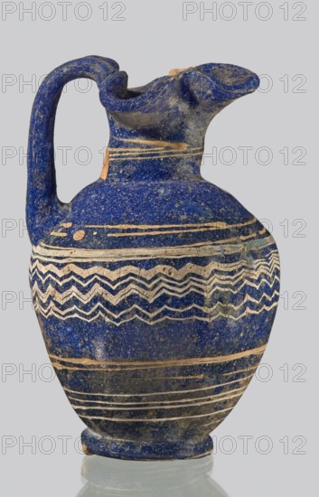 Oinochoe, unknown eastern workshop; 3rd century BC. Type: Grose group II:A, form II:3. Dark blue glass with threaded decoration on the belly of horizontal and zigzag lines in white and yellow glass widest point. Trefoil rim; molding on the core. Height: 8.90 cm; Body diameter: 5.60 cm; Weight: 0.076 kg., The image shows a single ceramic jug with a rounded body, narrow neck, flared spout, and a curved handle attached from the neck to the shoulder; the jug has a worn, speckled deep blue surface with bands and zigzag decorative patterns in a beige or sandy color, and areas of dark gray and lighter blue speckling; the handle and spout share the same blue with patches of beige wear, and the jug sits on a short foot that reflects slightly on the pale gray background.