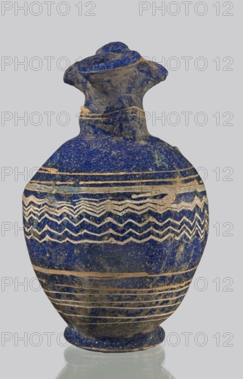 Oinochoe with a vertical arch-shaped handle; eastern workshop;, 3rd century BC. Type: Grose group II:A, form II:3. Dark blue glass with threaded decoration on the belly consisting of horizontal and zigzag lines in white and yellow glass at the widest point. Trefoil rim; molding on the core. Height: 8.90 cm. Body diameter: 5.60 cm. Outer diameter of rim: unknown. Outer diameter of the bottom: unknown. Vessel oinochoe. Weight: 0.076 kg., A rounded vessel with a narrow neck and a flared, ruffled rim sits upright; the surface is predominantly deep blue with a speckled texture and areas of darker and lighter blue, plus visible patches of beige and tan where the blue has worn away. The body of the vessel is decorated with a band of zigzag and wavy linear patterns in light beige contrasted against the blue, and several horizontal beige bands encircle the lower and upper portions. The neck and rim show rough, uneven surfaces with traces of tan and gray, and the vessel rests on a short circular...