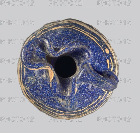 Oinochoe with a vertical arch-shaped vice; eastern workshop;, 3rd century BC Type: Grose group II:A, form II:3 Dark blue glass; on the belly threaded decoration in the form of horizontal and zigzag lines at the widest point of white and yellow glass. Trefoil rim; molding on the core Height: 8.90 cm Body diameter: 5.60 cm Outer diameter of the rim: ? cm Outer diameter of the bottom: ? cm Vessel, oinochoe, weight 0.076 kg, A top-down view of a rounded object with an irregular central opening, primarily deep blue with speckled lighter flecks, showing areas of worn tan or beige along raised edges and curves; the surface has flowing ridges and a few concentric bands in tan and blue, with smooth and textured patches and a neutral light gray background.