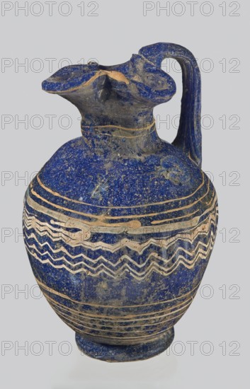 Oinochoe with a vertical arch-shaped vice; eastern workshop;, 3rd century BC. Type: Grose group II:A, form II:3. Dark blue glass with threaded decoration on the belly of horizontal and zigzag lines in white and yellow glass at the widest point. Trefoil rim; molding on the core. Height: 8.90 cm; body diameter: 5.60 cm; weight: 0.076 kg. Vessel oinochoe., A ceramic jug with a rounded body, narrow neck, flared and scalloped rim, and a curved handle attached from rim to shoulder; the surface shows a deep blue base color with areas of lighter blue and beige worn spots, and decorative bands of beige wavy and linear patterns encircling the body; the rim and handle show irregular edges and patches of beige where the blue coating is missing, and the overall texture appears rough and aged.