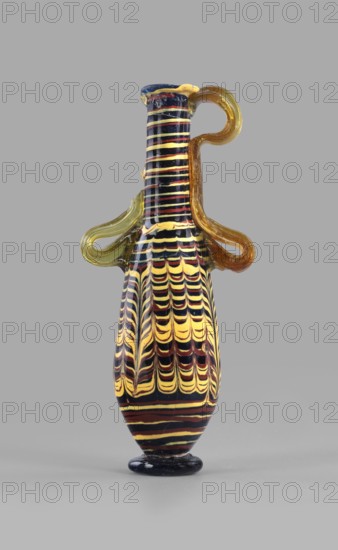 Amphoriskos; with an elongated neck; eastern Syrian or Cypriot workshop;, 2nd–1st century BC. Type: Grose group II:E, form III:4. Opaque glass with dark blue-green body and decoration of festoon patterns made with yellow and brown threads; discoidal rim; neck widening slightly downwards; baggy belly; solid foot. Vertical vices one partially chipped, double vices made of translucent light honey-green thread; formed on the core. Outer rim diameter 2.25 cm; height 14.80 cm; body diameter 4.10 cm; bottom diameter 2.85 cm. Vessel., A slender decorative vessel with a narrow neck and rounded body sits against a plain gray background; the vessel features alternating curved stripe patterns in yellow, dark blue, and reddish-brown, with the stripes forming a scalloped, feather-like motif on the body and horizontal bands on the neck. The vessel has a glossy finish and a small dark blue base, and it includes a translucent amber handle that loops from the neck to the shoulder and a second shorter tr...