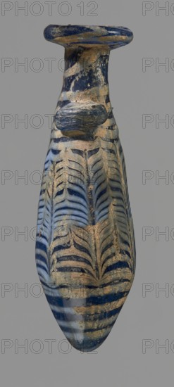 Alabastron with feathered decoration, Egyptian workshop, 6th–4th century BC, vessel alabastron, height 10.6 cm, width 3.7 cm, An elongated vessel with a narrow neck and a flared rim, displaying a marbled pattern of deep blue and pale blue with areas of tan or beige encrustation; the surface shows curved, featherlike repeating motifs in blue against lighter blue background, with several horizontal banded stripes toward the base and visible irregular patches of tan material and wear; the rim and neck have darker blue swirls and a rounded applied blob of glass, and the overall shape tapers to a rounded point at the bottom.