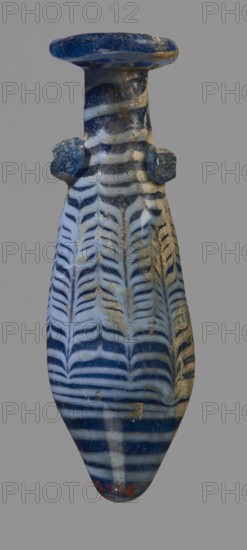 Alabastron with feathered decoration, Egyptian workshop, 6th–4th century BC, unknown place of origin; vessel alabastron, height 10.6 cm, width 3.7 cm., A narrow, elongated vase-shaped object with a flared rim and a long neck, showing a pattern of alternating dark blue and pale blue bands forming chevron-like feather shapes along the body; the piece has a glossy surface with areas of worn or rough texture and patches of sandy beige residue, and a pair of small rounded protrusions on opposite sides of the neck; the dominant colors visible are dark blue, pale blue, beige, and small hints of rusty orange.