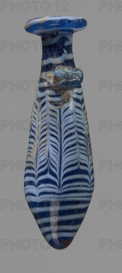Alabastron with feathered decoration, Egyptian workshop, 6th–4th century BC, vessel alabastron, height 10.6 cm, width 3.7 cm, A tall narrow glass vessel with a rounded lip and a tapered body, decorated with alternating bands and feather-like vertical patterns; the glass shows a deep blue base color with lighter blue and white streaks and swirls, and scattered areas of darker discoloration and surface roughness; a small rounded protrusion is attached near the neck, showing similar blue tones and surface wear; the background is a uniform gray.