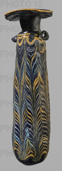 Alabastron, cylindrical belly, 4th–3rd century BC, vessel alabastron, height 17.4 cm, width 4.4 cm, origin unknown., A tall narrow vessel photographed against a neutral gray background, with a flattened rim and short neck leading to a long tapering body; the surface is predominantly dark, glossy black with decorative flowing featherlike patterns in yellow and light blue that run vertically along the body, and a band of undulating yellow lines near the shoulder; the rim and small looped handles show black with yellow edging, and the overall appearance is of a glazed, slightly worn object with fine linear motifs and subtle surface speckling.