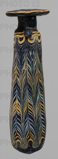 Alabastron, cylindrical belly, 4th–3rd century BC, vessel alabastron, height 17.4 cm, width 4.4 cm, A tall, narrow object with a rounded top and a flat disc-like cap sits against a plain gray background; the surface is predominantly dark blue or black with decorative flowing patterns in yellow and pale blue forming vertical feather-like or leaf-like motifs that repeat along its length, and a band of wavy yellow decoration just below the cap; the texture appears glossy and slightly worn in places, with a small protrusion beneath the cap and a rounded bottom showing continuation of the patterned design.