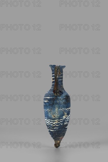 Amphorisko, Amphorisko with an ovoid belly, a long cylindrical neck ending in an uneven, slightly flaring rim, an uneven foot glued on, vertical, high, arched vises. Type: Grose III:E form III:2B, Harden form 7B. Opaque, dark blue glass decorated with white thread in a festoon pattern. The foot and vertical vises are made of dark amber, translucent glass; molding on the core. 2nd–1st century BC, Syria, vessel amphorisko. Rim diameter 2.8 cm, weight 0.110 kg., A narrow, elongated vase stands upright against a plain gray background, primarily showing deep blue tones with lighter blue areas and white wavy banding around its body; there are darker brownish vertical streaks near the neck and a small darker base with hints of brown and a tiny spot of reddish color; the surface shows some lighter, worn patches and thin horizontal striations, and the vessel has a single narrow neck that flares slightly at the rim.