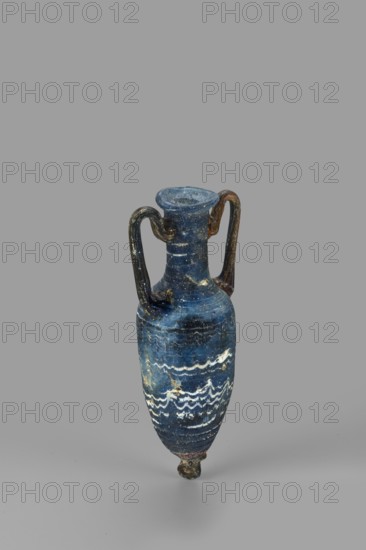 Amphorisko, Amphorisko with an ovoid belly, a long cylindrical neck ending in an uneven, slightly flaring rim, an uneven foot glued on, vertical high arched handles. Type: Grose III:E form III:2B, Harden form 7B. Opaque dark blue glass decorated with white thread in a festoon pattern. The foot and vertical handles are made of dark amber, translucent glass; molding on the core. 2nd–1st century BC, Syria; vessel amphorisko. Rim diameter 2.8 cm; weight 0.110 kg., The image shows a small, narrow amphora-like vessel with two curved handles attached near the neck, standing upright against a plain gray background. The vessel is predominantly deep blue with lighter blue variations and areas of wear showing off-white and pale yellowish patches; there are decorative wavy bands in white around the lower body. The handles appear darker, with brownish and black tones, and the base shows darker brown and slightly reddish traces. The rim and neck share the deep blue color with some lighter speckling ...