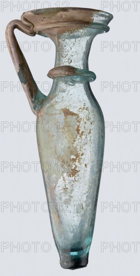Bottle; eastern workshop, 3rd–4th century AD. Transparent bluish-green glass, free-blown, with short cylindrical neck and funnel-shaped rim; two applied glass threads of same color in a ring beneath rim. Outer rim diameter 5.20 cm; height 15 cm; belly diameter 5.30 cm; bottom diameter 1.65 cm; weight 0.066 kg., A slender, elongated glass vessel with a narrow base widening into a rounded body and a flared rim, featuring a curved handle attached from the rim to the shoulder and a decorative glass ring around the neck; the surface shows weathered, patchy areas and encrustations with a worn texture. The visible colors are pale blue-green glass, areas of brown and tan encrustation, whitish and grayish weathering, and some darker greenish-blue at the base.