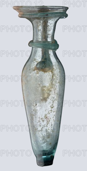 Bottle; eastern workshop;, 3rd–4th century AD. Transparent bluish-green glass, short cylindrical neck, funnel-shaped rim with two applied glass threads in a ring beneath the rim; free-blown. Outer rim diameter 5.20 cm. Height 15 cm. Belly diameter 5.30 cm. Bottom diameter 1.65 cm. Vessel, height 15 cm, width 5.3 cm, weight 0.066 kg., A single elongated glass vessel with a narrow neck and flared rim, shown upright against a plain light gray background; the glass appears translucent with a greenish-blue tint and areas of cloudy white and pale brown discoloration across its surface; there is a rounded glass collar encircling the neck and a thicker ring of glass around the rim, both showing the same green-blue color; the body tapers smoothly toward a slightly rounded base that has a deeper green-blue hue and darker irregular spots; the surface shows irregular textured patches, streaks, and small surface imperfections and inclusions.