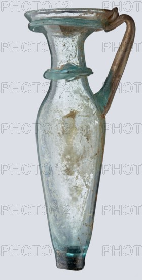 Bottle; eastern workshop, 3rd–4th century AD. Transparent bluish-green glass, free-blown; short cylindrical neck and funnel-shaped rim with two applied glass threads in a ring beneath the rim. Outer rim diameter 5.20 cm; height 15 cm; belly diameter 5.30 cm; bottom diameter 1.65 cm; weight 0.066 kg. Unknown place of origin., A tall translucent glass vessel with a narrow elongated body that tapers toward the base and flares at the rim, showing a smooth curved handle attached from near the rim down the shoulder; the glass appears weathered with a cloudy patina and surface encrustation, and there is a raised decorative band around the neck; visible colors include pale aqua, light blue, greenish tints, brownish spots, and patches of white on the glass surface.