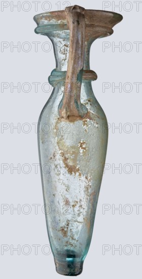 Bottle; eastern workshop, 3rd–4th century AD. Transparent bluish-green glass, free-blown, with a short cylindrical neck and a funnel-shaped rim. Two applied glass threads of the same color form a ring under the rim. Outer rim diameter 5.20 cm; height 15 cm; belly diameter 5.30 cm; bottom diameter 1.65 cm; weight 0.066 kg., A tall narrow glass vessel with a single curved handle attached from the neck to the shoulder, showing a weathered surface with patches of encrustation and rough textured areas; the glass is mostly a pale aqua to light blue-green with areas of cloudy white and pale beige deposits, and there are rusty brown and tan discolorations scattered across the body and neck; the rim flares outward and appears thicker and slightly irregular, and the handle has streaks of muted brown and green with some flaky buildup; the base is rounded with a darker teal hint and shows worn and roughened areas.