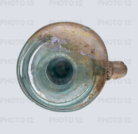 Bottle; eastern workshop, 3rd–4th century AD. Transparent bluish-green glass, free-blown; short cylindrical neck and funnel-shaped rim with two applied glass threads in a ring beneath the rim. Outer diameter of rim: 5.20 cm. Height: 15 cm. Belly diameter: 5.30 cm. Bottom diameter: 1.65 cm. Vessel; height 15 cm; width 5.3 cm; weight 0.066 kg., A circular glass vessel viewed from above with a rounded rim and a short handle on one side, showing a mix of greenish-blue and pale turquoise glass tones, with areas of brownish tan and iridescent purplish sheen across the upper surface, patches of dull green and muted yellowish discoloration, and some cloudy and weathered textures on the interior and rim.