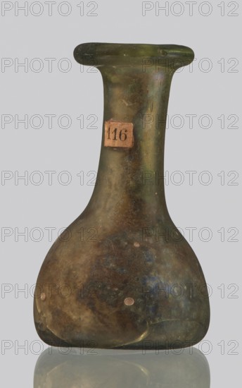 Vial, unknown eastern workshop, hemispherical body with a flat bottom, high cylindrical neck with an annular spout. 3rd–4th century AD, vessel, height 4 cm, width 1.3 cm, A rounded vessel with a narrow neck and flared rim, shown against a pale gray background and reflected on a glossy surface, with an overall mottled surface in shades of green, olive, brown, and hints of blue, along with a few small pale spots and areas of darker discoloration.