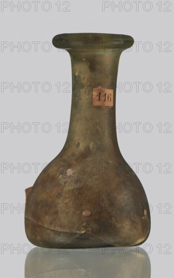 Vial, unknown eastern workshop, hemispherical body with a flat bottom, high cylindrical neck with an annular spout. 3rd–4th century AD. Unknown place of origin. Vessel; height 4 cm, width 1.3 cm., A worn glass bottle with an elongated neck and flared rim, showing a rounded shoulder that tapers to a broad base; the surface has a cloudy, uneven texture with patches of discoloration and iridescent sheen in shades of green, brown, and olive, with traces of darker and lighter areas and small spots of a pale tan color; the bottle sits on a reflective surface, and the background is a uniform light gray.