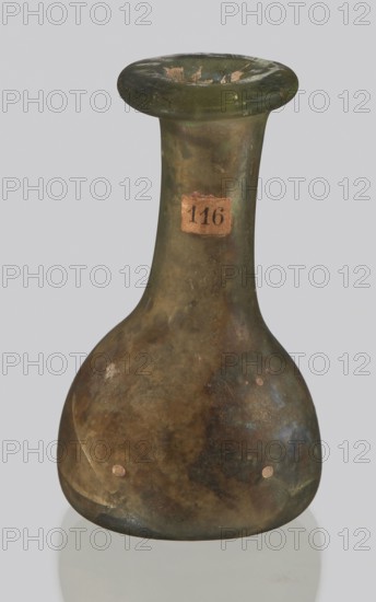 Vial, unknown eastern workshop, hemispherical body with a flat bottom, high cylindrical neck with an annular spout. 3rd–4th century AD, vessel; height 4 cm, width 1.3 cm, A greenish-brown glass bottle with a rounded base and a long narrow neck topped by a flared rim, showing surface iridescence and patches of darker brown and green tones with some lighter worn spots, a small rectangular label affixed to the neck, and the bottle resting on a reflective pale gray surface against a light gray background.